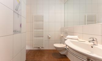A modern bathroom with white tiles and a large mirror. It is well-lit and tidy, featuring a sink and a toilet.