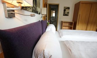 A cozy hotel room with a comfortable bed and modern lighting. The walls are light and the furniture is simple and functional.