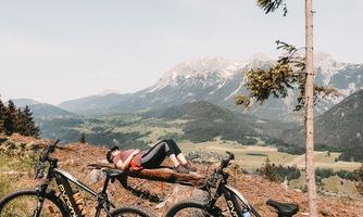 Two mountain bikes are standing in a meadow with a view of the mountains. In the background, a person is relaxed on a lounge chair.