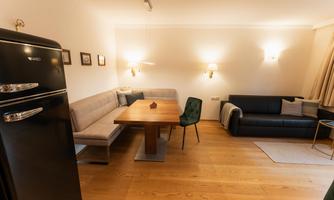 A cozy living room with a large table, a comfortable bench, and a refrigerator. The walls are painted in neutral colors, and there is soft lighting.
