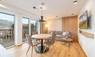 A modern living room with wooden floors and large windows. A round table with chairs is in the room, and a cozy couch invites relaxation.
