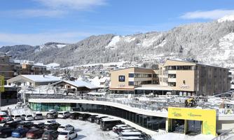 A winter cityscape with snow-covered mountains in the background. Parking lots and modern buildings are visible in the scene.