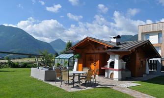 A cozy garden house with a table and chairs is surrounded by green meadows and mountains in the background. The sky is clear with a few clouds.