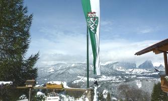 A winter landscape with snow-covered mountains in the background. In the foreground, a flagpole with a green and white flag.