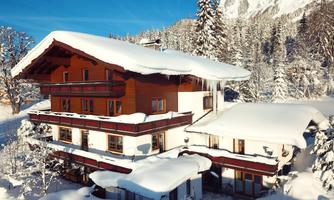 A cozy chalet in the snow, surrounded by snow-covered trees and majestic mountains. The clear blue sky complements the wintry landscape.
