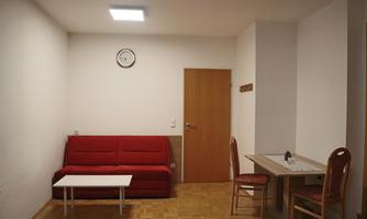 A simple living room with a red sofa and a small table. A clock is hanging on the wall, and there is a chair as well as a table on the right side.