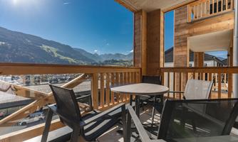 A sunny balcony with a table and chairs, surrounded by impressive mountains. The sky is clear and blue, perfect for relaxing moments outdoors.