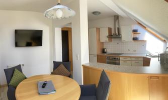 A cozy dining area with a round table and modern chairs. The open kitchen is well-equipped and has plenty of natural light.