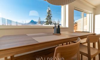 A bright and inviting dining area with a wooden table. Through large windows, the snow-covered landscape and the clear sky are visible.