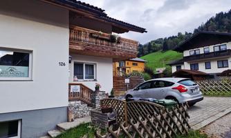 A modern house with a balcony in a picturesque setting. In the foreground, there is a car, and in the background, green hills and other buildings can be seen.