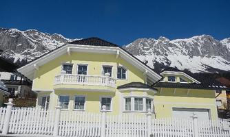 A bright yellow house with a balcony stands in the foreground. Impressive mountains and a clear, blue sky can be seen in the background.