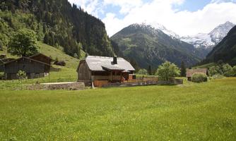 An idyllic old wooden house in a green meadow, surrounded by mountains. The sky is blue with some clouds.