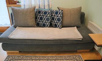 A cozy sofa with two large cushions and a soft throw. Next to it is a small table on a beautiful rug.