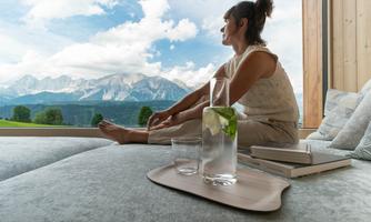 A woman sits relaxed in the room and looks at the mountains through the window. Next to her on a table is a glass of water with lime.