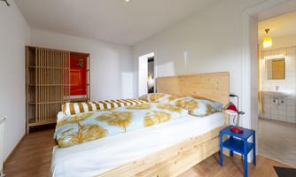 A cozy bedroom with a large bed and bright blankets. In the background, there is a wardrobe and access to a bathroom.