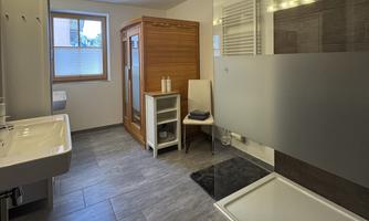 A modern bathroom with a shower, a sink, and a wooden sauna. The room is bright and friendly with large windows.