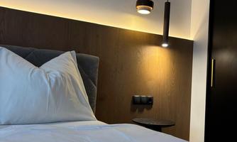 A modern bedroom with a cozy bed and elegant lighting. The walls are in warm wood and gray tones, creating a relaxing atmosphere.