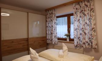 A cozy bedroom with a large window and floral curtains. Neat pillows and blankets lie on the bed.