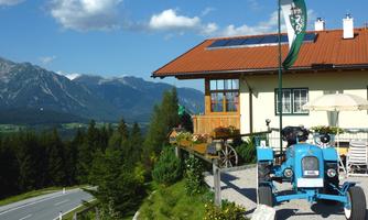 A picturesque country house with a blue tractor in front. In the background, mountains and a clear sky can be seen.