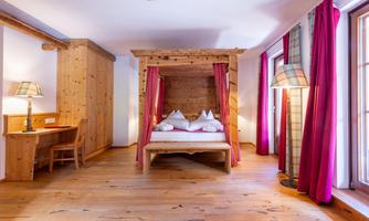 A cozy bedroom with a four-poster bed and wooden floor. The walls are decorated with red curtains and there is a small desk area.