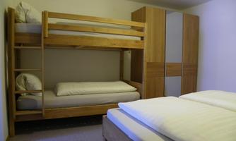 A simple bedroom with a bunk bed and a single bed. There are wooden wardrobes and a calm atmosphere.