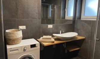A modern bathroom with dark tiles. It includes a washing machine, a stylish sink, and wooden shelves.