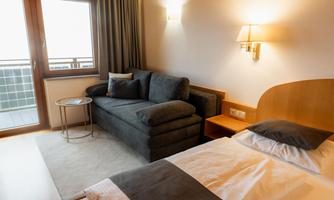 A cozy hotel room with a double bed and a gray couch. There is a small table and large windows with access to a balcony.