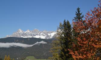 An impressive mountain landscape with snow-covered peaks and a clear blue sky. In the foreground, autumn trees shine in warm colors.