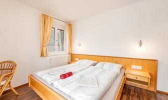 A cozy bedroom with a double bed and wooden floor. The room is bright and has a window with curtains.