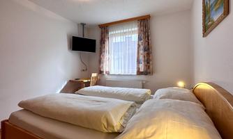 A bright guest room with two single beds and a television on the wall. Natural light enters the room through the window.