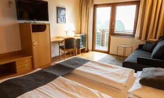 A cozy hotel room with two beds, a television, and a table. Large windows provide a view of the balcony and the outdoor area.