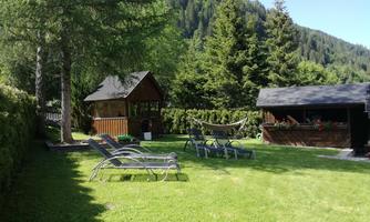 A green meadow with deck chairs and a wooden house. In the background, tall trees and a mountain landscape can be seen.