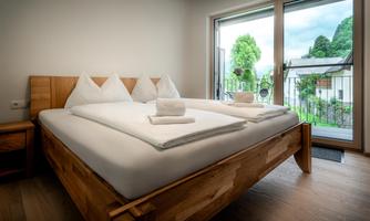 A modern bedroom with a large double bed and fresh towels. Large windows offer a view of the green surroundings.