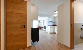 A modern hallway with a wooden door and a window to the kitchen. The floor is laid with light wood and the walls are in neutral colors.