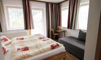 A cozy room with a double bed and a sofa bed. There are large windows with curtains and a small table.