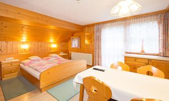A cozy room with wooden walls, a double bed, and a dining area. Large windows let in plenty of light, creating an inviting atmosphere.