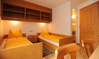 A simple bedroom with two beds and a wooden shelf wall. The cushions are in a bright yellow and the floor is covered with a patterned carpet.