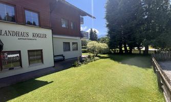 A beautiful green meadow in front of a wooden building named "Landhaus Kogler". In the background, trees and a clear sky can be seen.