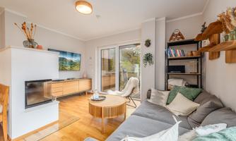 A modern living room with a cozy couch and a round table. Large windows let in plenty of light and provide access to a balcony.