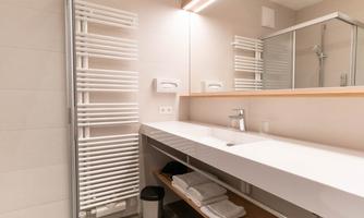 A modern bathroom with a simple vanity and a large mirror. Towels and a waste bin are neatly stored.
