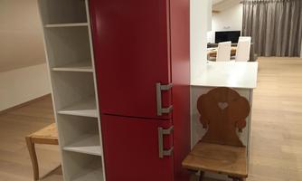 A modern kitchen with a red refrigerator and a small wooden chair. In the background, shelves and a dining area can be seen.