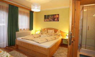 A cozy bedroom with a large wooden bed and green curtains. In the background, there is a bathroom with a glass shower.