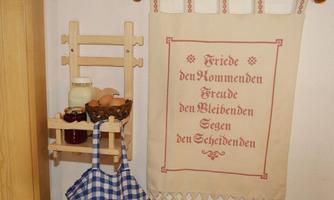 A decorative wall tapestry with the inscription "Peace to those who come, joy to those who stay, blessings to those who depart." Below it is a wooden shelf with eggs, a glass of milk, and jam.