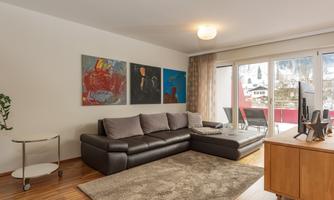 A modern living space with a large, dark gray sofa and colorful pictures on the wall. Large windows offer a view of the surroundings and let in a lot of light.