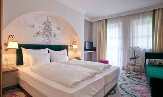 An elegant hotel room with a double bed and a detailed mural. The room is bright and cozy, featuring a modern armchair and plants.