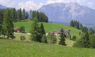 A picturesque landscape with green meadows and wooded hills. In the background, majestic mountains rise under a clear sky.