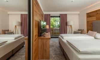 A modern bedroom with two beds and a television. Large windows let in plenty of daylight and offer a view of nature.