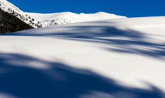 A snow-covered landscape under a clear blue sky. The gentle hills cast long shadows in the snow.
