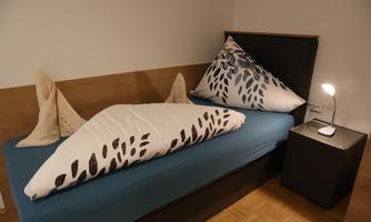 A simple bed with blue bedding and decorative pillows in the shape of fish fins. Next to the bed is a nightstand with a modern lamp.