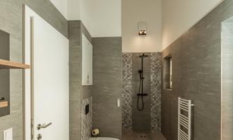 A modern bathroom with a shower and a toilet. The walls are in neutral gray tones and there are large windows that provide plenty of light.